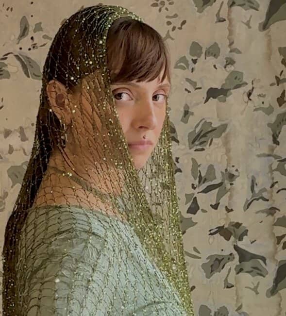 Close-up of the gold mesh veil framing Renata's face against painted wallpaper.