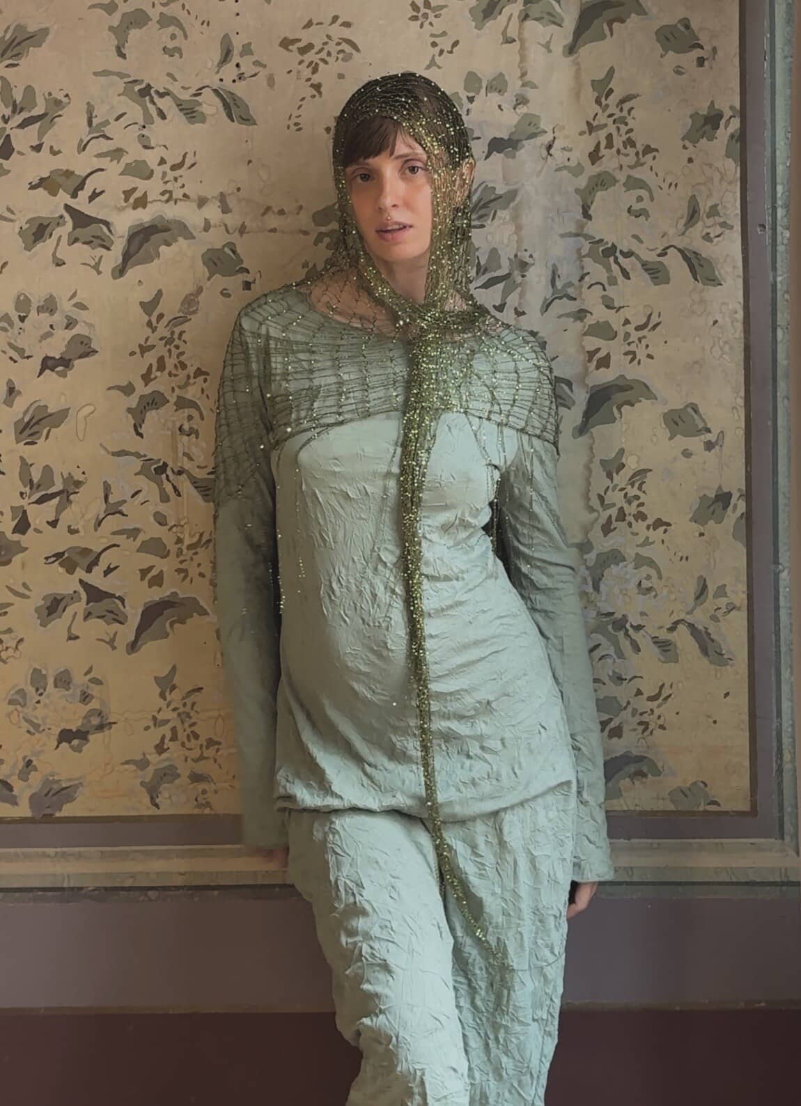 Renata leaning on painted wallpaper, gold mesh draped as a hood and shoulder cape with long tails.