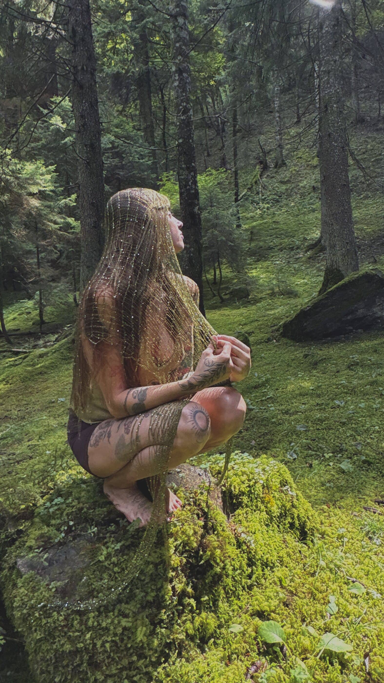 Renata seated on a mossy boulder in a pine forest, face turned up into a shaft of sunlight, a gold mesh veil falling down her back.