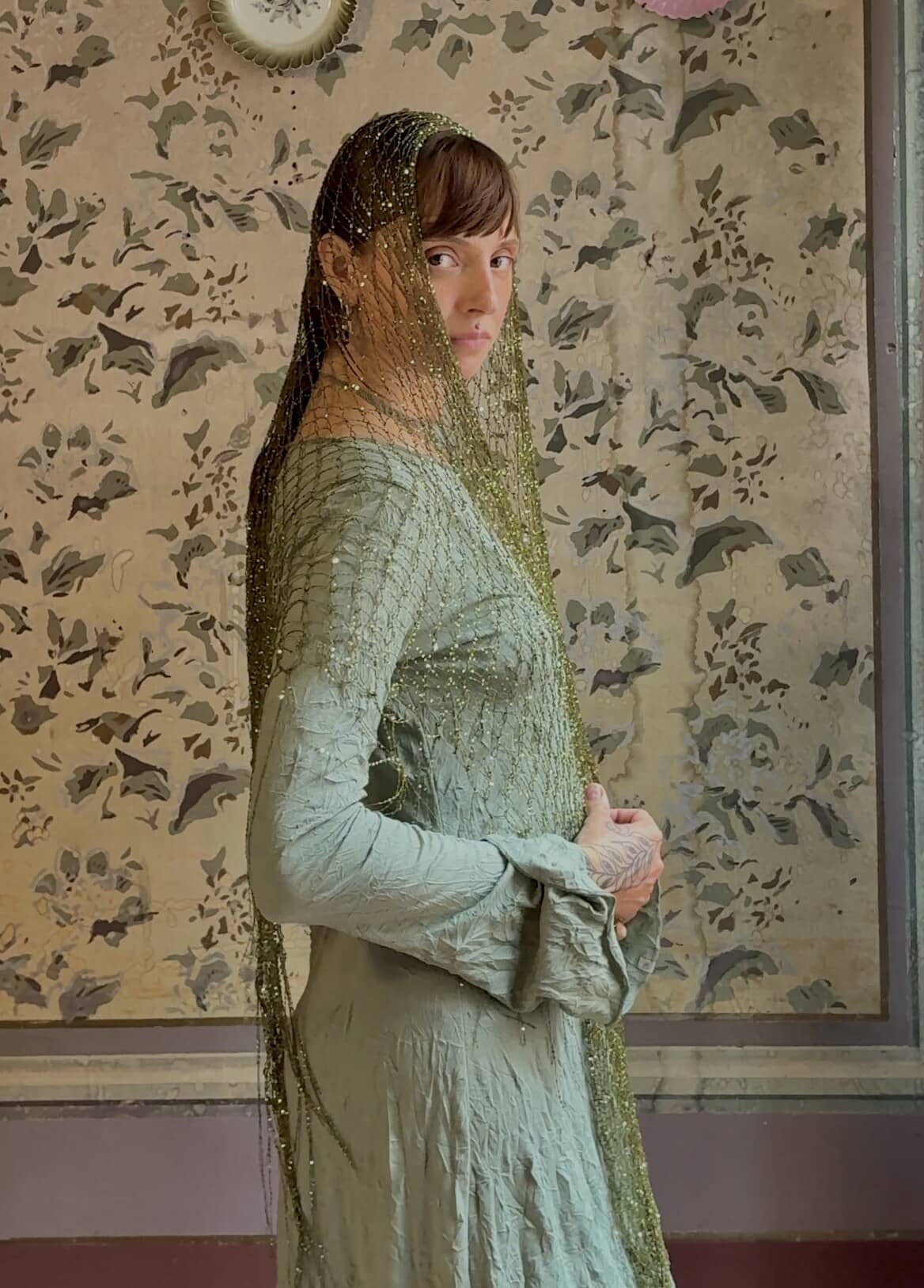 Renata in profile against faded hand-painted floral wallpaper, a gold mesh veil over her head and a sage silk dress.