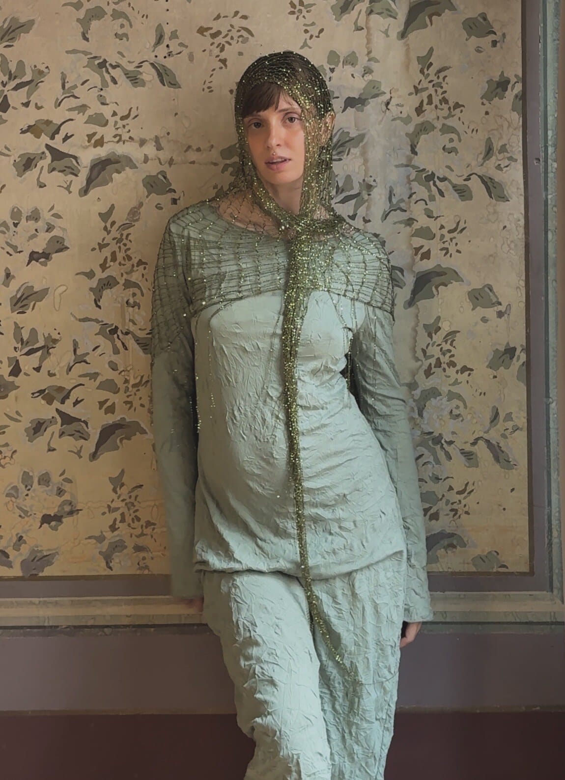 Renata leaning on painted wallpaper, gold mesh draped as a hood and shoulder cape with long tails.