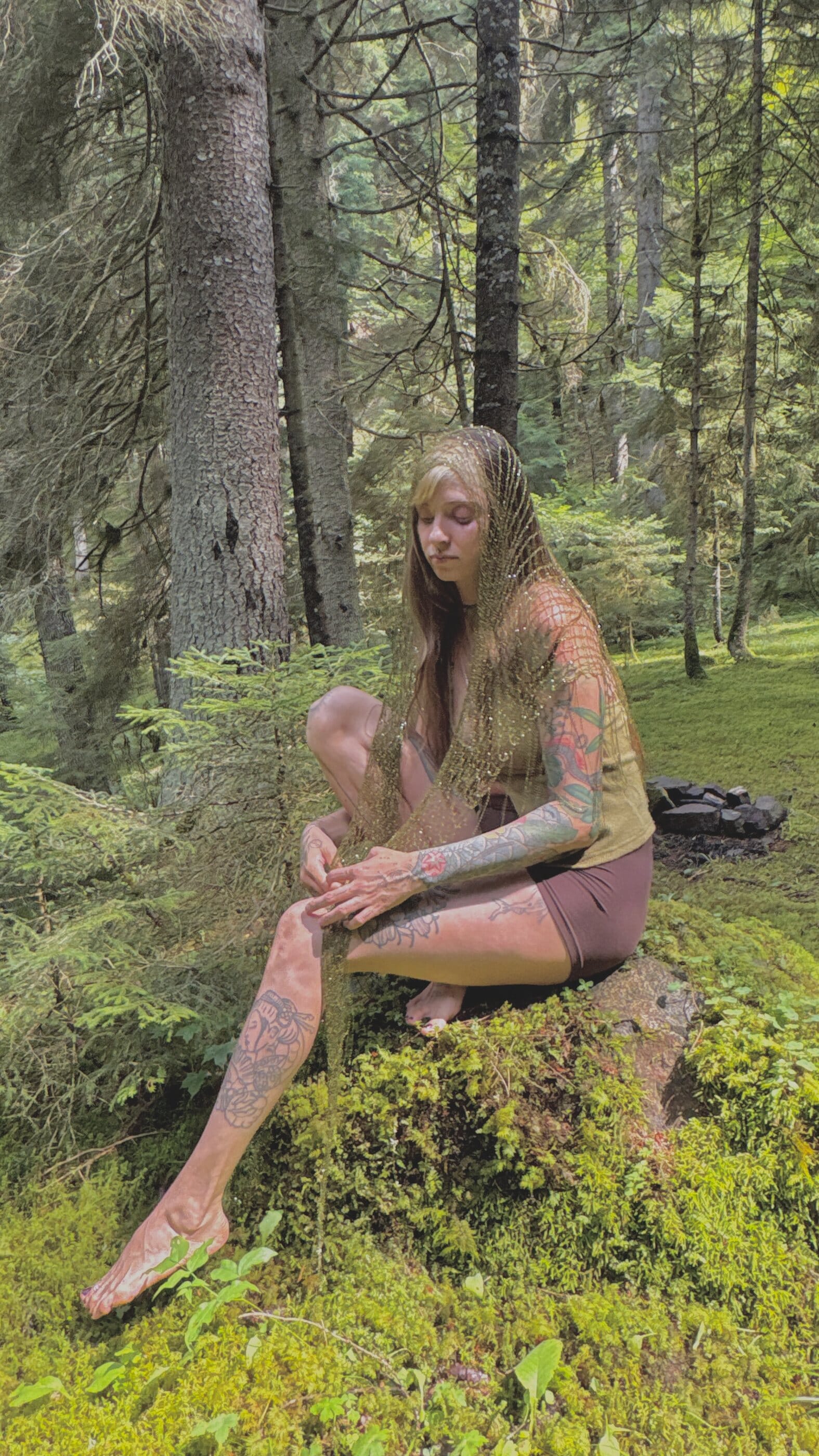 Renata sitting on moss beneath tall pines, looking down at the gold mesh in her hands.