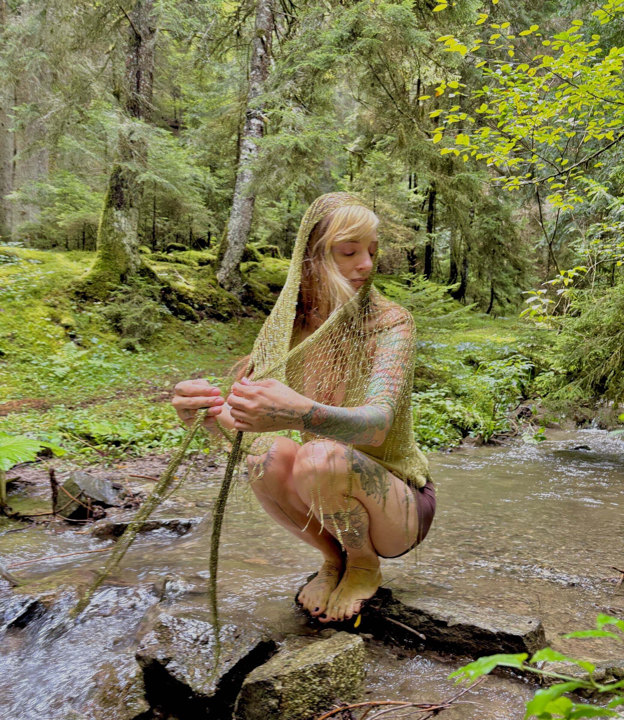 Renata crouched on stones in a forest stream, hooded in gold mesh, holding its long knotted tails over the water.